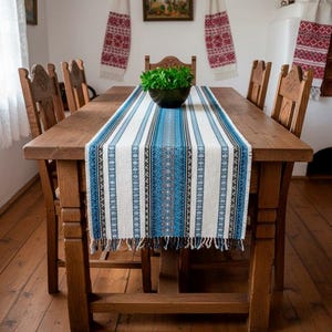 May include: A wooden dining table with six wooden chairs. A blue and white patterned table runner with fringe detail runs down the center of the table. A dark green bowl with greenery sits on the table runner. Decorative wall hangings are in the background.