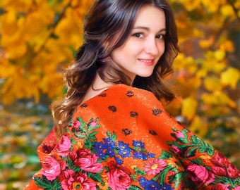 May include: A woman with long brown hair is wearing a red shawl with a floral pattern. The shawl has a bright orange background with pink, blue, and green flowers. The woman is standing in front of a background of yellow leaves.