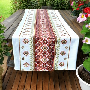 Puede incluir: Un camino de mesa blanco con un patrón bordado en rojo, amarillo y verde. El camino de mesa está sobre una mesa de madera con flores en macetas a cada lado.