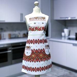 Op de afbeelding: Witte schort met rood en zwart geborduurde bloemen- en geometrische patronen. De schort heeft een rij rode rozen en twee zakken. Het ontwerp wordt getoond op een mannequin voor een vage keukenachtergrond.