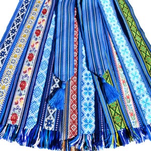 Puede incluir: Una colección de cinturones textiles azules tejidos intrincadamente con varios patrones y colores. Algunos cinturones presentan diseños florales, mientras que otros muestran formas geométricas. Cada cinturón tiene una borla en el extremo y los bordes están deshilachados.