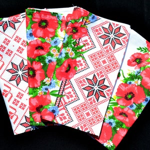 May include: Four white fabric napkins with red poppy floral and geometric cross-stitch patterns. The napkins feature vibrant red poppies, blue cornflowers, and green leaves. The cross-stitch designs include red and black starburst and diamond patterns.