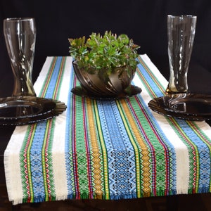 May include: A table runner with a colourful striped pattern in blue, green, yellow, and red. The runner is on a dark table, with a plant in a brown bowl, two tall glasses, and two plates. The runner has a fringed edge.