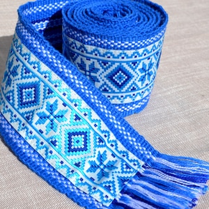 May include: A blue woven ribbon with a white and blue geometric pattern and fringe. The ribbon is rolled up and has a tassel on one end.