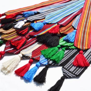 Puede incluir: Una colección de cintas tejidas de colores con borlas. Las cintas están hechas de diferentes colores y patrones, incluyendo rojo, azul, verde, negro y blanco. Las borlas están hechas del mismo material que las cintas y están unidas a los extremos.