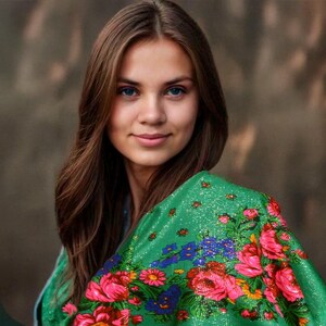 May include: A young woman with long brown hair is wearing a green shawl with a floral pattern. The shawl has pink, red, blue, and yellow flowers on it.