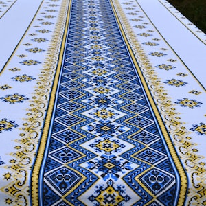 May include: White tablecloth with a blue and yellow embroidered pattern. The pattern features geometric shapes and floral motifs.
