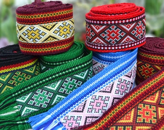 May include: A selection of colourful woven ribbons and rolled fabric. The ribbons display intricate geometric patterns in various colours, including red, green, blue, and yellow. The fabric rolls are in shades of red and burgundy.