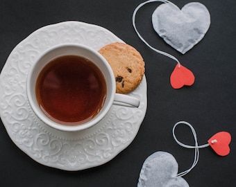 May include: A white teacup and saucer with a cookie on a black background. There are four heart-shaped tea bags with strings hanging from them.