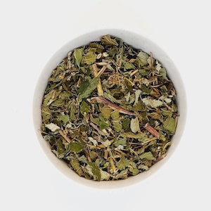 May include: A white bowl filled with dried green and brown herbs. The herbs are loose and appear to be a blend of different types.