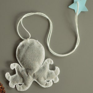 May include: An octopus-shaped tea bag made from a light, semi-transparent material. The tea bag has eight tentacles and is attached to a white string. A small, pale blue star is attached to the string. The background is a solid, muted grey.