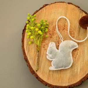 May include: A white tea bag shaped like a squirrel on a wooden slice with a brown pom-pom and a sprig of yellow flowers.