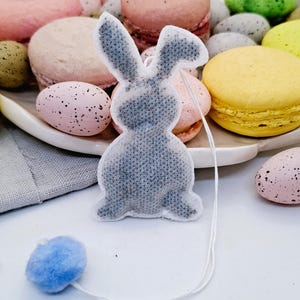 Könnte beinhalten: Ein grauer, hasenförmiger Teebeutel mit einem blauen Bommel. Er liegt vor einem Teller mit pastellfarbenen Makronen und gesprenkelten Ostereiern und schafft eine festliche, frühlingshafte Präsentation.