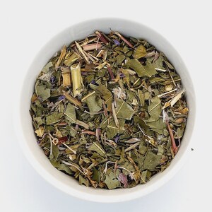 May include: A white bowl filled with a blend of dried herbs and flowers, including green leaves, brown stems, and purple flower petals.