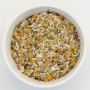 May include: A white ceramic bowl filled with dried chamomile flowers. The flowers are a mix of yellow and white, with a few brown stems. The bowl is set against a plain white background, highlighting the natural colors of the tea.