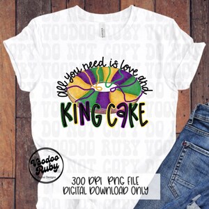 以下が含まれることがあります： カラフルなキングケーキのデザインと「all you need is love and KING CAKE」の文字が入った白いTシャツ。緑、紫、金色の要素が含まれており、マルディグラのお祝いに最適です。