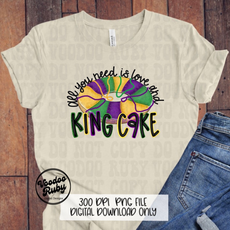 以下が含まれることがあります： クリーム色のTシャツ。キングケーキのグラフィックと「All you need is love and King Cake」のテキストが特徴です。デザインには緑、紫、金色の要素が含まれており、マルディグラをテーマにしています。シャツはブルージーンズと組み合わせています。