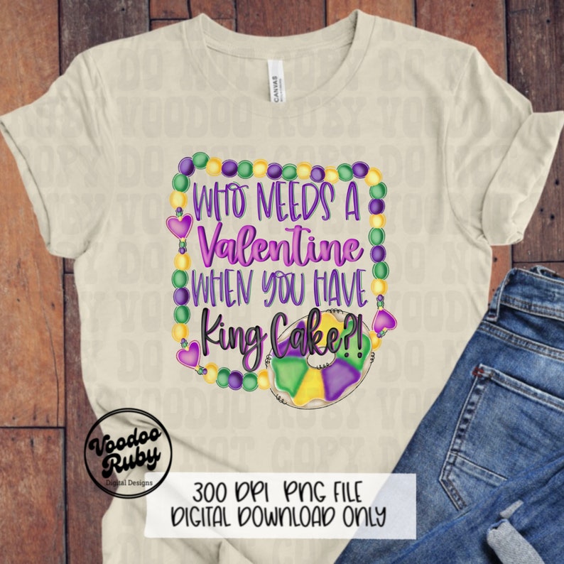 May include: A cream-colored short-sleeve t-shirt featuring a graphic design with purple and yellow text that reads Who Needs A Valentine When You Have King Cake?! The design is framed by purple, green, and yellow beads and a king cake illustration.