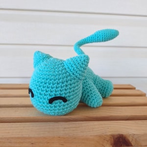 Può includere: Un giocattolo a forma di gatto all'uncinetto turchese con occhi neri e una lunga coda. L'animale di peluche fatto a mano è appoggiato su una superficie di legno. Il gatto ha orecchie a punta e un corpo arrotondato.