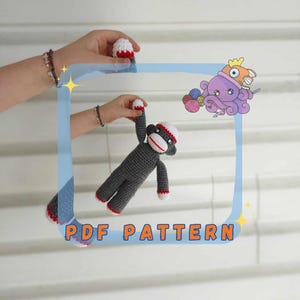 May include: A crochet pattern for a gray and red sock monkey. The pattern is available as a PDF download. The image shows a person holding the finished sock monkey.