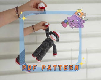 May include: A crochet pattern for a gray and red sock monkey. The pattern is available as a PDF download. The image shows a person holding the finished sock monkey.