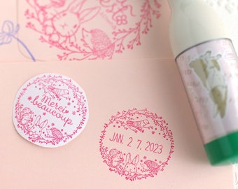 May include: A pink rubber stamp with a floral wreath design featuring a squirrel, rabbit, and hedgehog. The stamp also includes the text "JAN. 2 7. 2023".