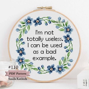 May include: A cross-stitch pattern with a floral wreath surrounding the text "I'm not totally useless. I can be used as a bad example." The pattern is in a wooden embroidery hoop and has a blue and green color scheme. The text "#110 PDF Pattern SmileXstitch" is below the hoop.