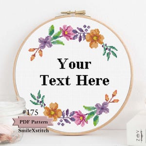 May include: A cross-stitch pattern featuring a floral wreath with purple, orange, and pink flowers. The wreath surrounds the text "Your Text Here". The pattern is labeled with the number "#175" and the text "PDF Pattern SmileXStitch".
