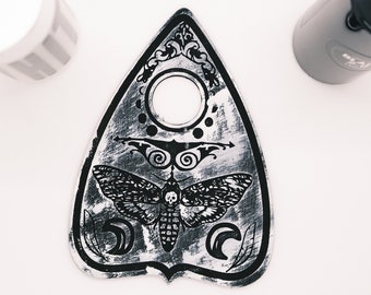 May include: A black and white ouija board with a moth design and a hole in the center. The board is made of wood and has a distressed finish.