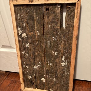 Puede incluir: Una pieza de arte mural rectangular de madera. La obra de arte está construida con tablones de madera recuperada, mostrando una apariencia rústica y desgastada. El marco es de madera de color claro, y los tablones son de color marrón oscuro con acentos blancos y verdes.