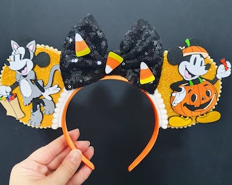 May include: Halloween headband featuring Mickey and Minnie Mouse. The headband has a black sequined bow with candy corn decorations, orange ears, and character cutouts on each side. The characters are set against a gold glitter background.