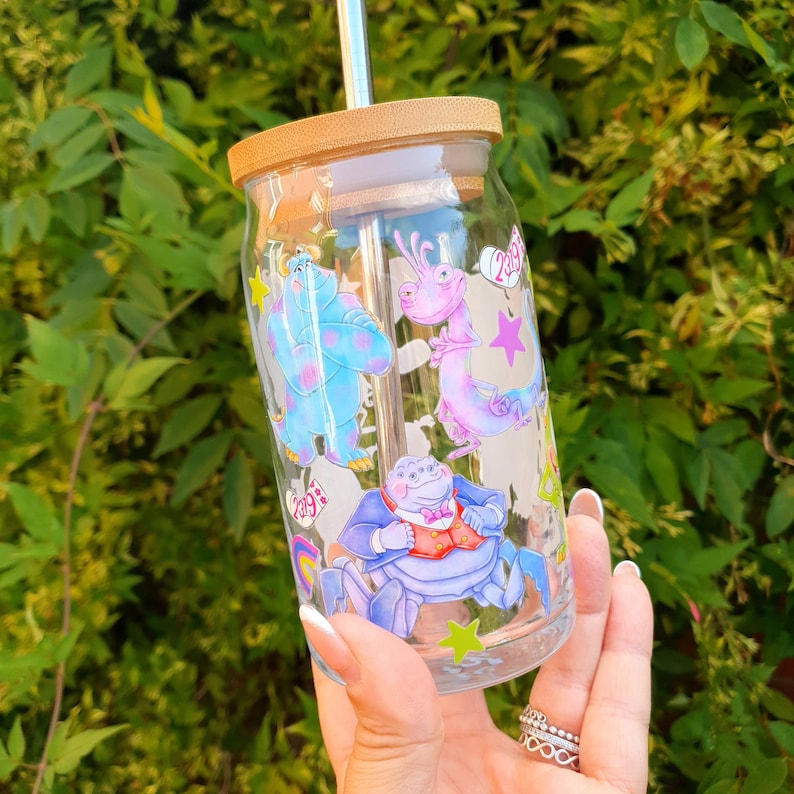 May include: A clear glass reusable water bottle with a bamboo lid and a straw. The bottle has a colorful design featuring characters from the movie Monsters Inc. The characters include Sulley, Mike, and Boo.