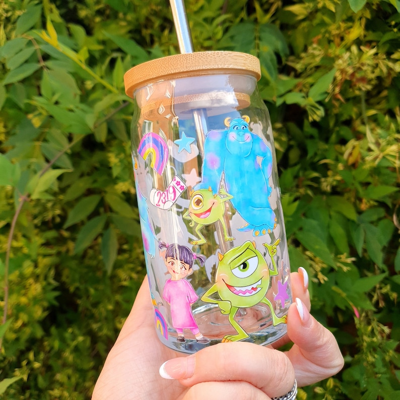 May include: A clear glass reusable water bottle with a bamboo lid and a metal straw. The bottle has a colorful design featuring characters from the movie Monsters Inc. The characters include Sully, Mike, Boo, and Randall.