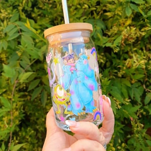 May include: A clear glass jar with a bamboo lid and a stainless steel straw. The jar has a colorful design featuring characters from the movie Monsters Inc. The characters include Sulley, Mike, and Boo.
