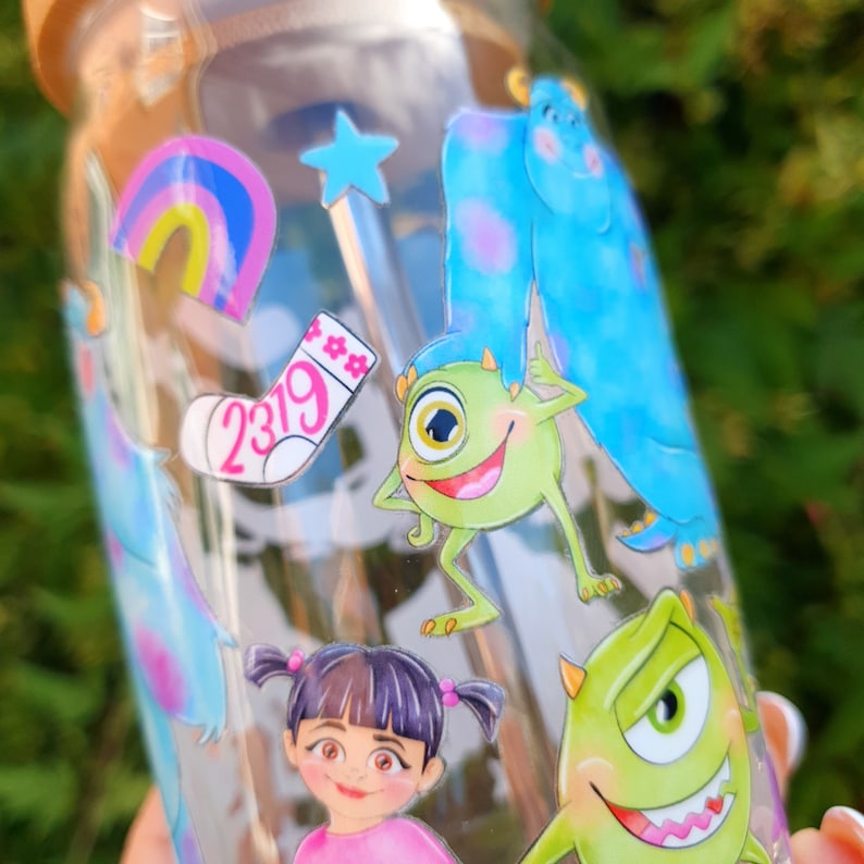 May include: A clear glass bottle with a cork lid decorated with colorful cartoon stickers. The stickers feature characters from the movie Monsters Inc., including Sully, Mike, and Boo. The bottle also has a rainbow sticker and a sock sticker with the number 2319.