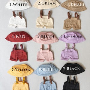 Può includere: Nove set di abbigliamento per bambini, ciascuno con un cappello da pescatore e una salopette abbinati. I colori includono bianco, crema, kaki, rosso, rosa, lavanda, giallo, blu e nero. I cappelli e le salopette hanno cinghie marroni.