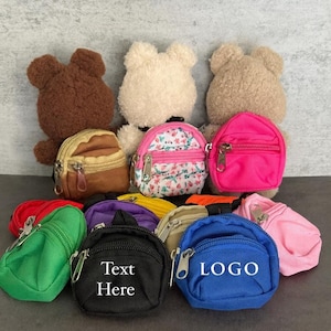 May include: A selection of miniature backpacks in a variety of colours, including pink, green, blue, and black, each with a zip closure. Some have the text "Text Here" or "LOGO". Three teddy bear heads are visible in the background.