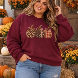May include: A maroon long-sleeve sweatshirt featuring a graphic print of three pumpkins in the centre. The pumpkins display distinct patterns including leopard print, brown polka dots, and a red and white plaid design, each topped with a bow.