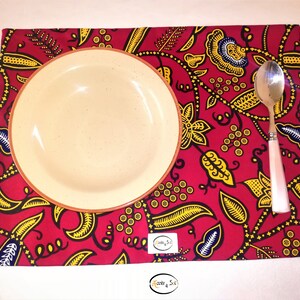 Peut inclure: Un set de table rouge avec un motif floral jaune et bleu, une assiette crème et une cuillère avec un manche rose. Le set de table porte une étiquette avec le texte "Roots & S.I."