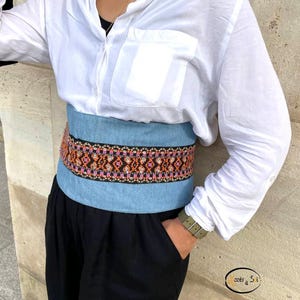 Peut inclure: Une ceinture en jean bleu clair avec un motif brodé coloré en orange, rose et noir. La ceinture est portée sur une chemise blanche et un pantalon noir. La ceinture mesure environ 10 cm de large.