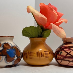 Puede incluir: Tres pequeños jarrones de cerámica con diferentes diseños pintados. El jarrón del medio es de color marrón claro con líneas marrones pintadas. El jarrón de la izquierda es blanco con diseños florales marrones y azules. El jarrón de la derecha es rosa claro con líneas marrones y rosa oscuro pintadas.
