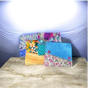 May include: A collection of handmade fabric wallets in various floral and patterned designs. The wallets feature a mix of colors, including blue, pink, yellow, and green, with button closures. The wallets are displayed on a marble-like surface.