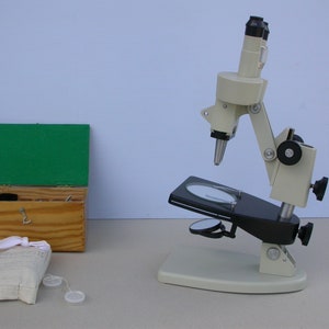 May include: A vintage microscope in cream and black. Beside it is a wooden box with a green lid, a white cloth bag, and two clear lens covers. The setup suggests a science or educational theme. The microscope is likely used for scientific study.