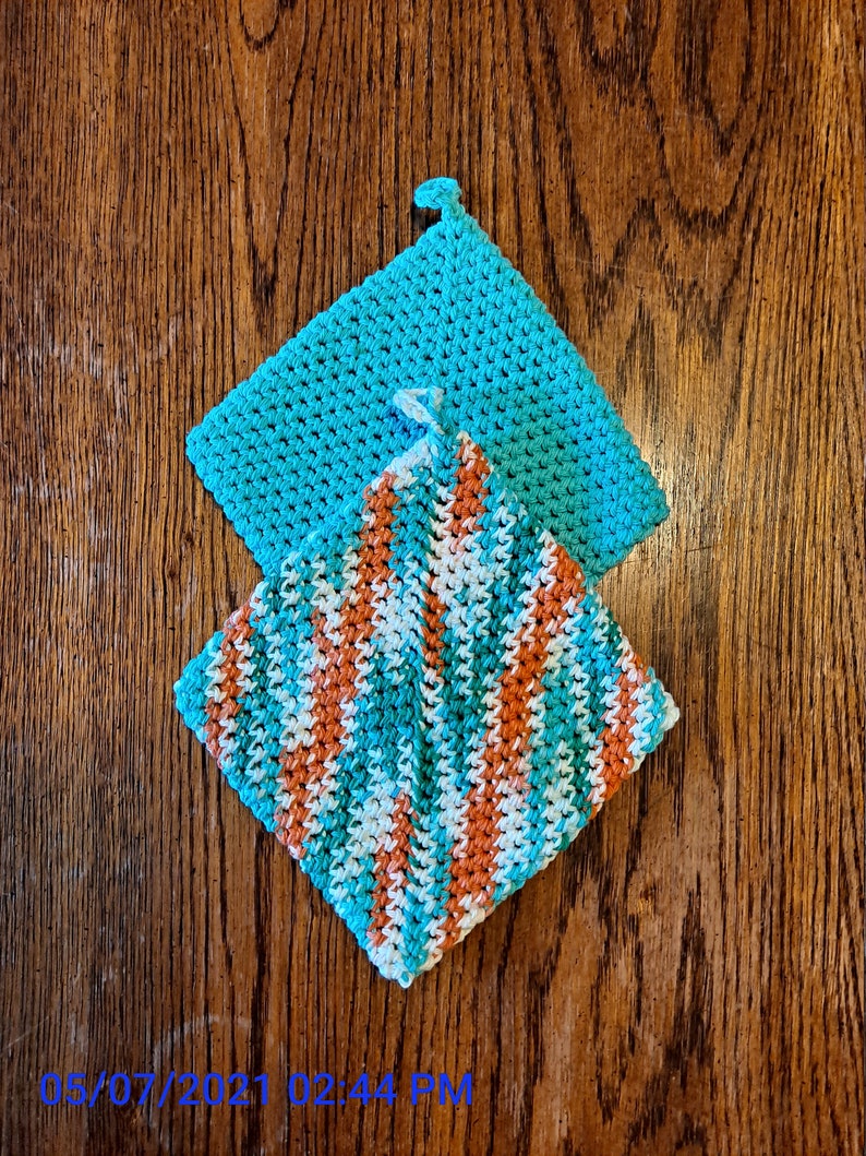 以下が含まれることがあります： 2つの編まれた鍋つかみ。1つはターコイズブルーで、もう1つはターコイズブルー、白、オレンジのストライプです。