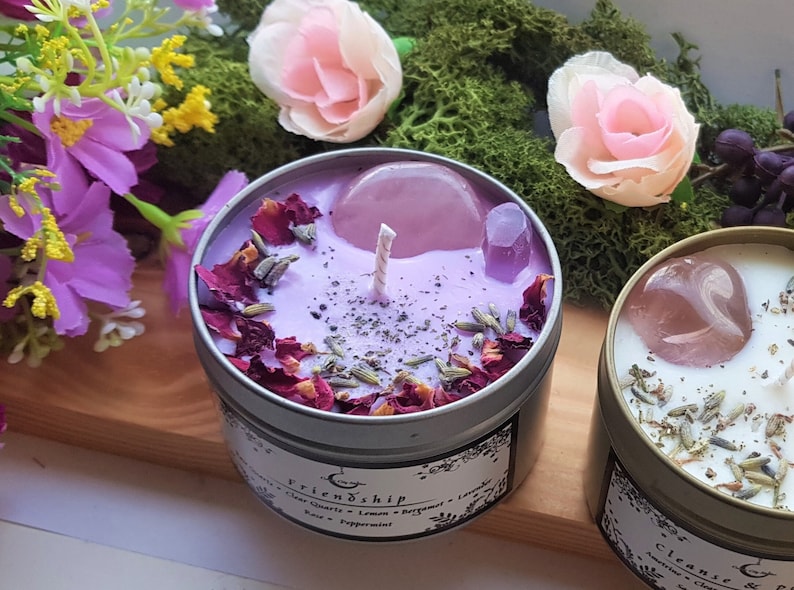 A smallish candle with pinkish/purple wax in a silver tin. On top are a tumbled rose quartz, a small clear quartz point, red rose petals, dried lavender, dried peppermint set into the wax in a circular pattern around the edge