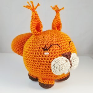 Puede incluir: Un juguete de peluche de ardilla naranja tejido a crochet con patas marrones y una nariz blanca. La ardilla tiene una sonrisa cosida y los ojos cerrados.