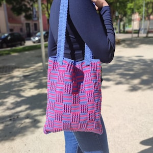Puede incluir: Una bolsa de mano de crochet azul y rosa con un patrón de cuadros. La bolsa tiene dos asas largas.