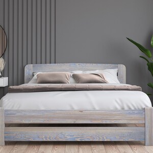 May include: A white wooden bed frame with a light blue wash finish. The bed has a headboard and footboard with a simple design. The bed is made of solid wood and has a natural wood grain. The bed is dressed with white bedding and two brown throw pillows. The bed is in a bedroom with a grey wall and a round mirror.