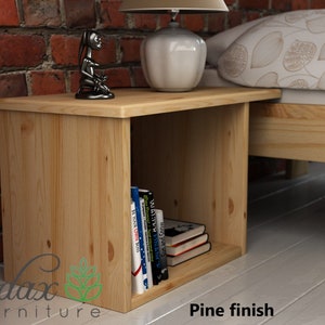 May include: A wooden bedside table with a shelf for books. The table is made of pine wood and has a natural finish. The table is next to a bed with a white and brown patterned bedspread.