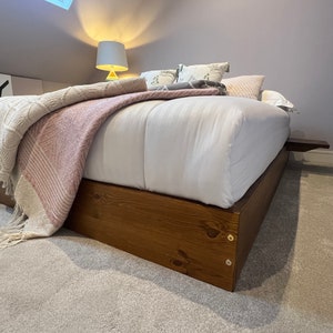May include: A wooden platform bed frame with a white mattress and pink and white blanket. The bed is made up with pillows and a throw blanket. The bed frame is stained a dark brown colour.